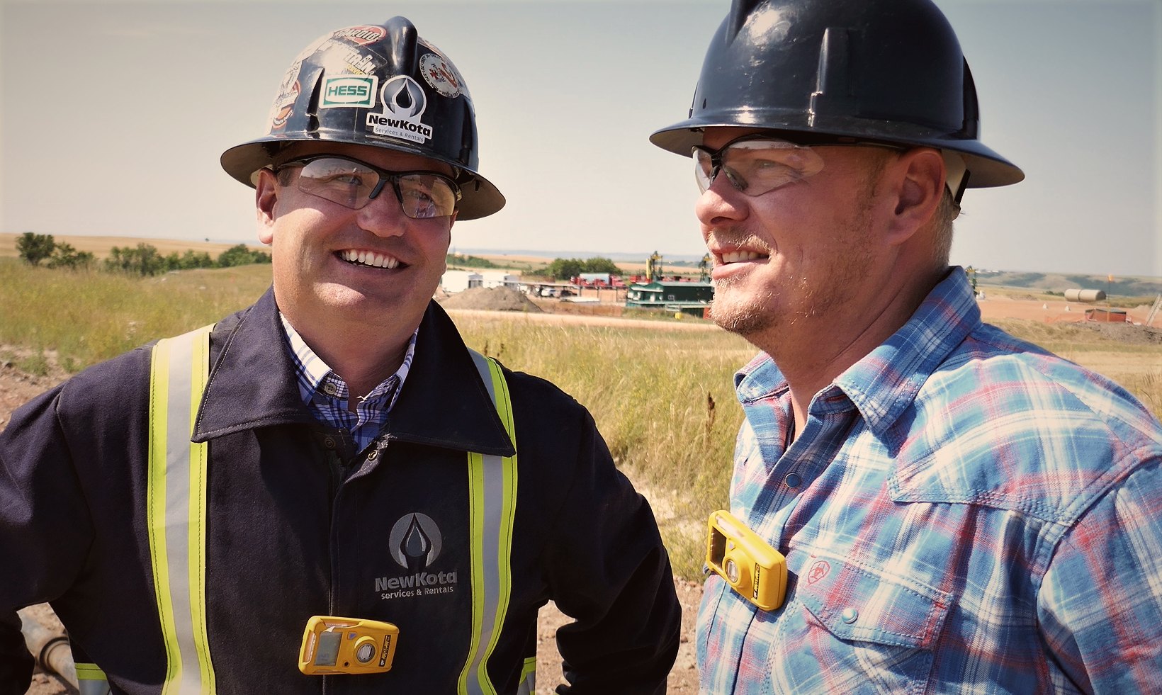 Two smiling men in hard hats and safety glasses on a North Dakota well site, one in a NewKota Services & Rentals jacket